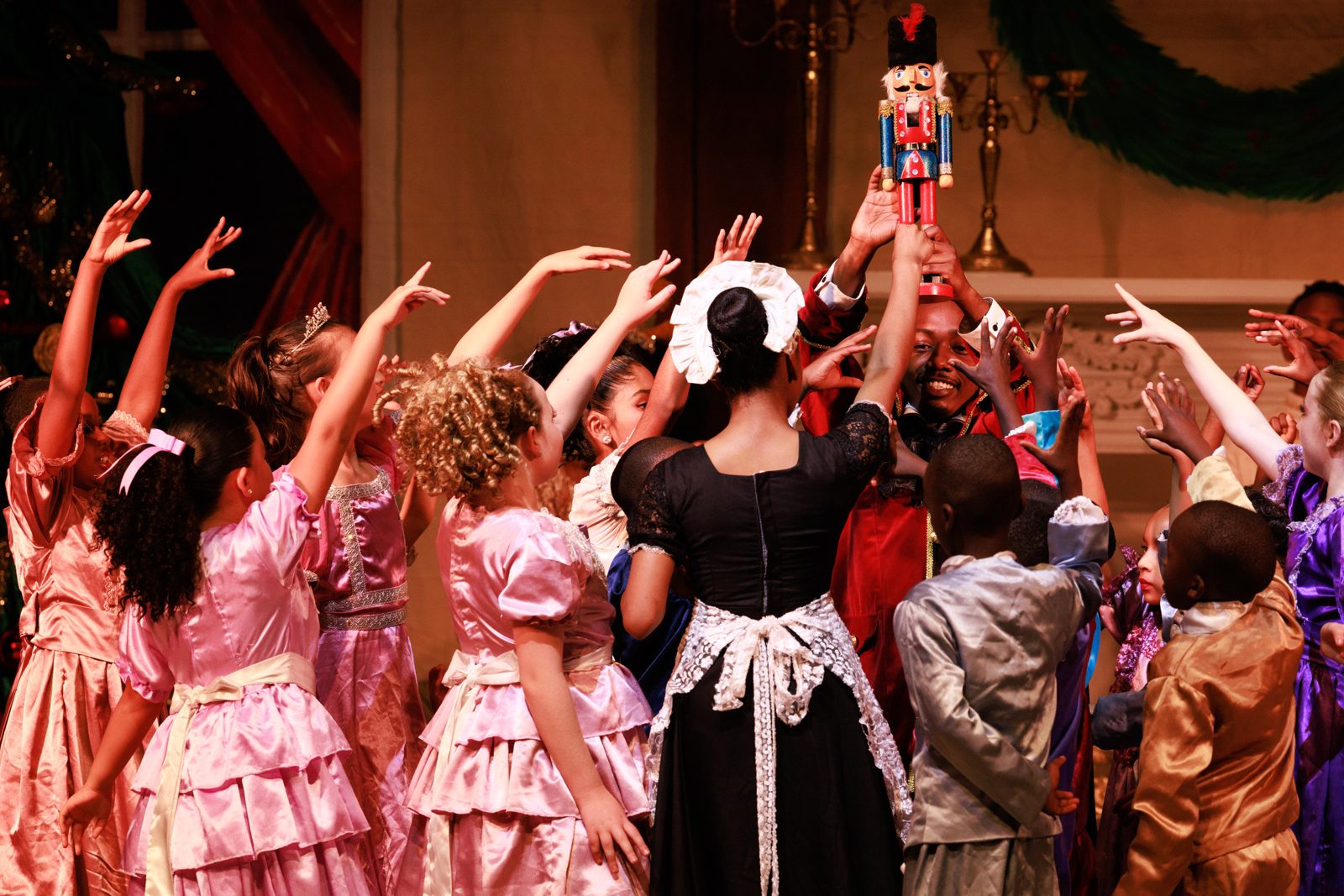 Children in party dresses reach up toward the nutcracker doll held high at the Christmas party