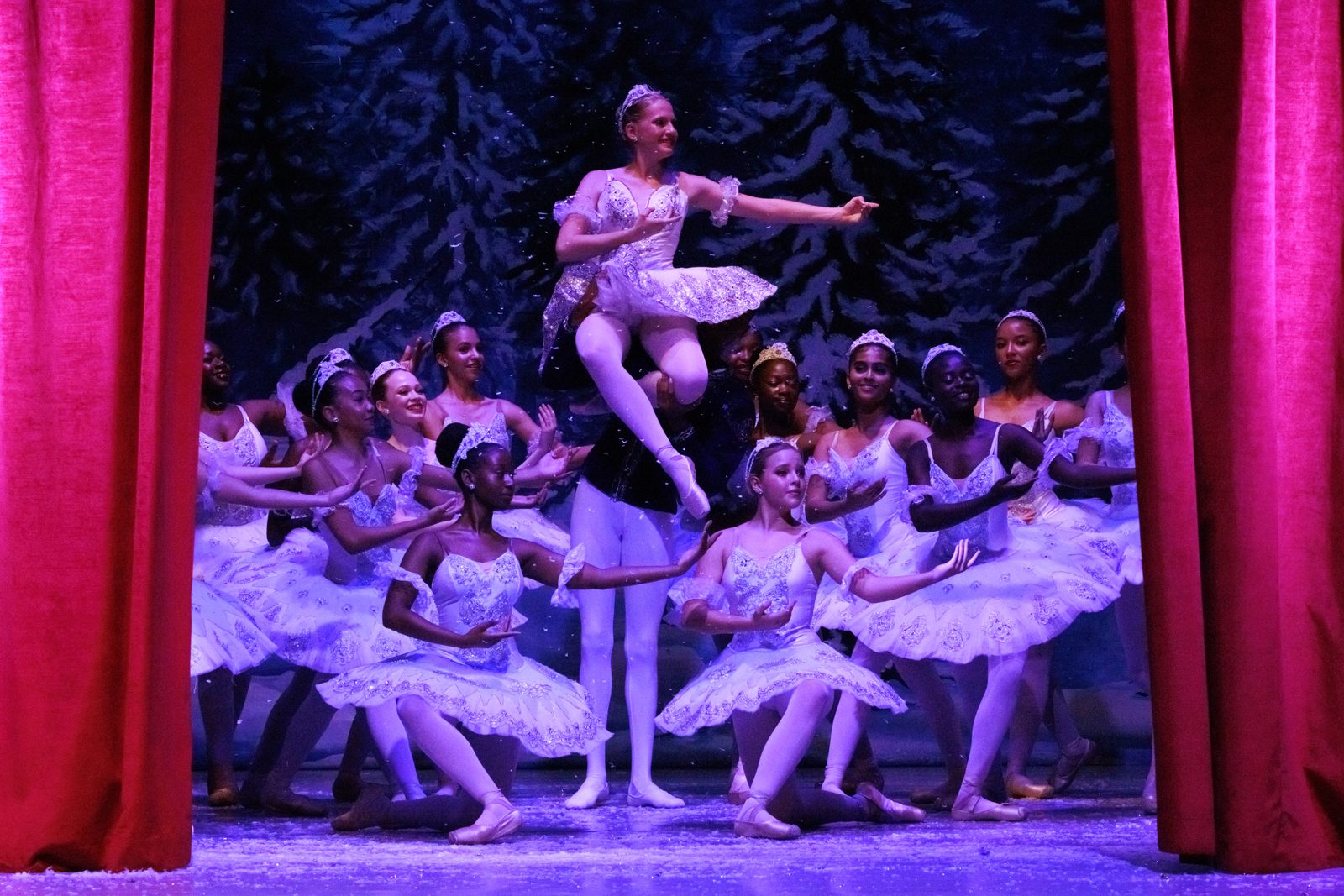The Snow Queen lifted above a circle of snowflake dancers in white tutus, framed by red stage curtains
